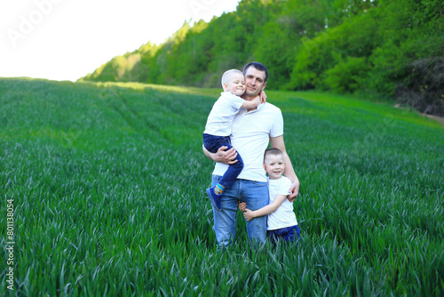 A man holding two young boys in a field of vibrant green grass. The man is dressed in a blue shirt and has a warm smile on his face, radiating happiness and contentment. The boys, who appear to be