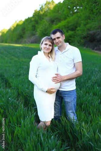 A couple standing in a beautiful field. The woman is wearing a white dress and has her hands gently placed on her stomach, indicating that she is pregnant. The man stands beside her, with his arm