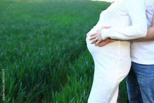 A couple standing in a beautiful field. The woman is wearing a white dress and has her hands gently placed on her stomach, indicating that she is pregnant. The man stands beside her, with his arm