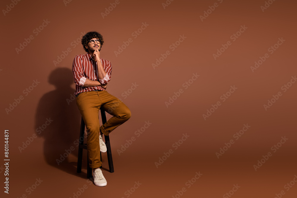 Fototapeta premium Full size photo of nice young man sit stool look empty space wear striped shirt isolated on brown color background