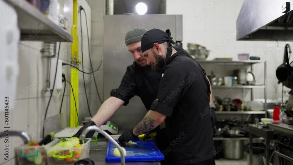 Two professional chefs in black uniforms work on preparations and cut vegetables while working in a restaurant kitchen. A male cook chops an onion while his colleague takes the chopped and prepared Two professional chefs in black uniforms work on preparations and cut vegetables while working in a restaurant kitchen. A male cook chops an onion while his colleague takes the chopped and prepared