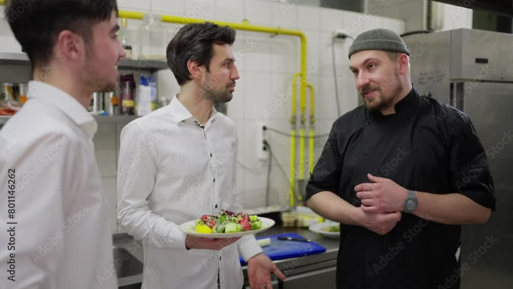 Communication between two male waiters in white shirts and a professional cook in a black uniform while solving professional issues in the kitchen of a restaurant Communication between two male waiters in white shirts and a professional cook in a black uniform while solving professional issues in the kitchen of a restaurant