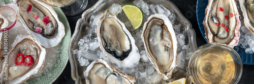 Oysters on ice panorama, with various toppings and white wine, overhead flat lay shot on a black stone background
