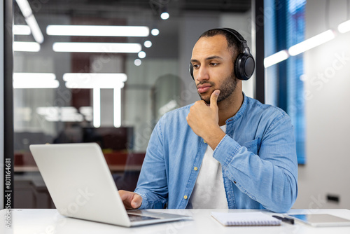 Thoughtful and serious Arab man, office worker, student working and studying in modern office wearing headphones and looking at laptop monitor screen