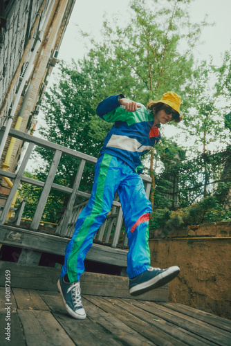 Young boy wearing vibrant '90s inspired sportswear in blue color, yellow panama and casual sport shoes, walking outdoors. Concept of 90s, fashion, youth culture, old-style trends