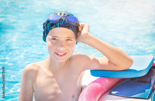 Happy child (boy) in cap, sport goggles ready to learns professional swimming with pool board, swim noodles and fins for swimming. Kid enjoying water in swimming pool. Healthy lifestyle.