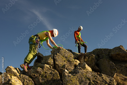 Two people are climbing a mountain, one of them wearing a green harness. The other person is wearing a red jacket