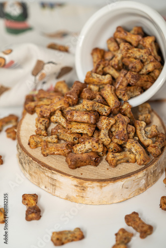 An assortment of gourmet dog treats shaped like bones, spilled from a ceramic bowl onto a wooden base