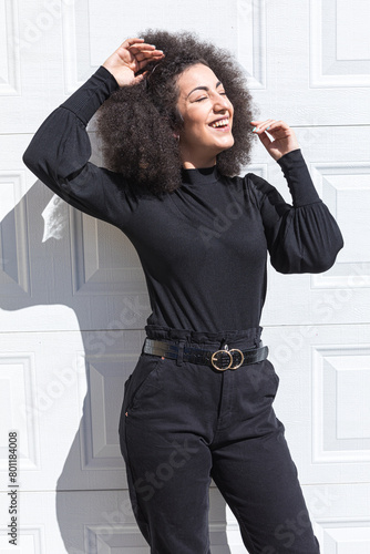 Young white woman with afro-style hair, dressed in black, leaning against a white door, smiling, wearing a headband, facing the sun.