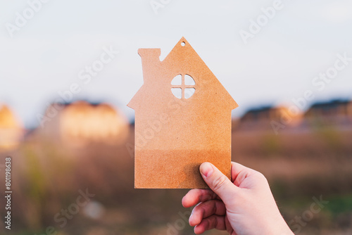 Man's hand holds a wooden house on the background of blurred buildings. Blurred outline of residential buildings in the background, suggesting themes of home, real estate, and community.
