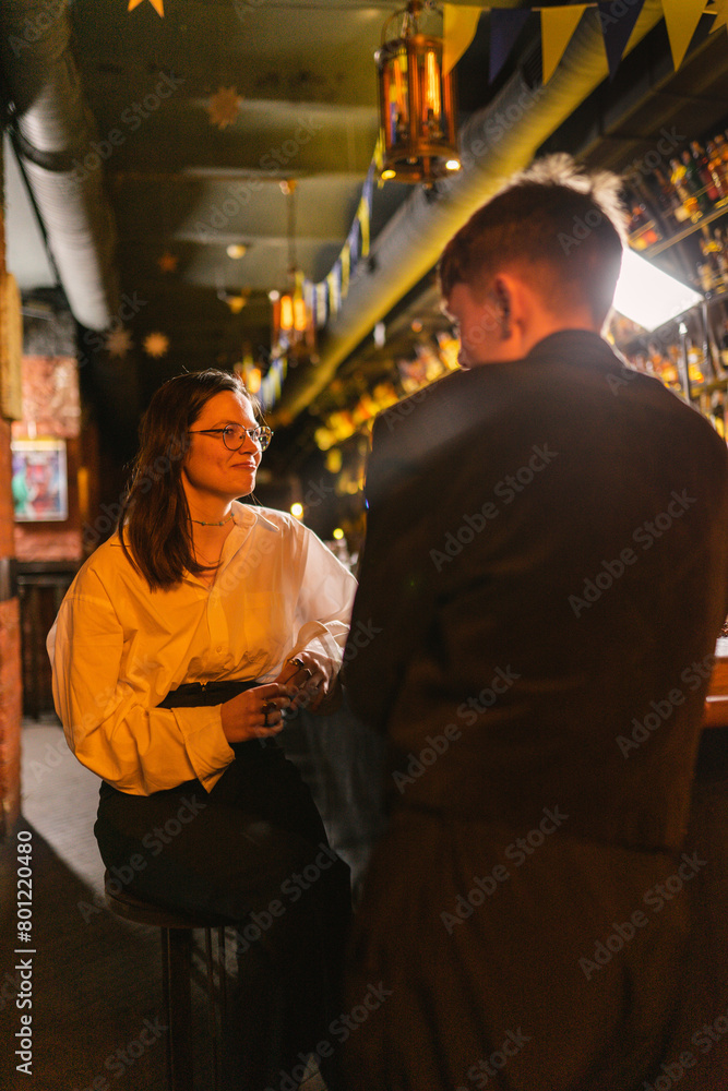 Fototapeta premium Smiling woman with eyeglasses looks at man in office suit in pub. Lady meets interesting friend at party on fancy night club