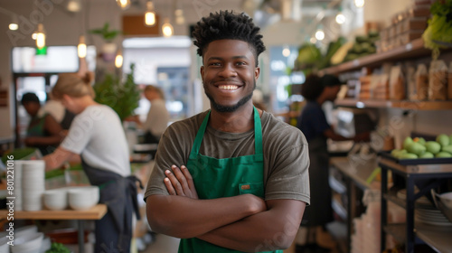 A cheerful young man in a green apron, arms crossed, oversees his eco-friendly store bustling with activity. photo
