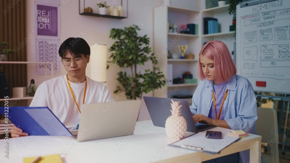 Asian game developer and his pink-haired colleague working on laptops in office Asian game developer and his pink-haired colleague working on laptops in office