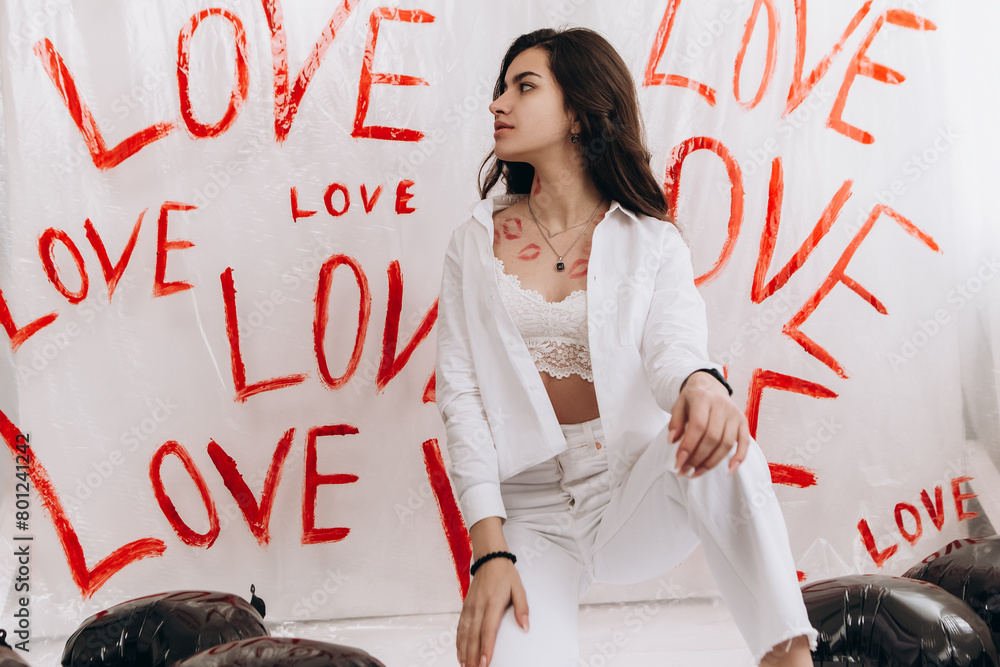 Fototapeta premium Woman sitting with kisses lipstick near black foil balloons, the word LOVE hand-painted on plain white surface oilcloth, Valentine`s day concept celebration decoration. Love, holiday, party 