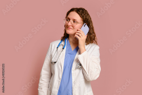 Woman doctor holding a mobile phone in her hands, studio pink background. Nurse in uniform with stethoscope on red studio background © Андрей Журавлев