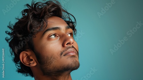 A young Indian man with his gaze thoughtful, against a solid teal background