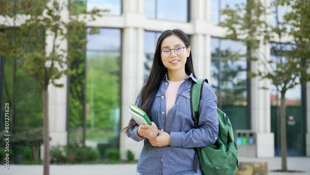 Portrait of smiling asian female student in glasses with books in hands standing on campus space near university building. Head shot of happy young woman applicant with backpack posing looking camera Portrait of smiling asian female student in glasses with books in hands standing on campus space near university building. Head shot of happy young woman applicant with backpack posing looking camera