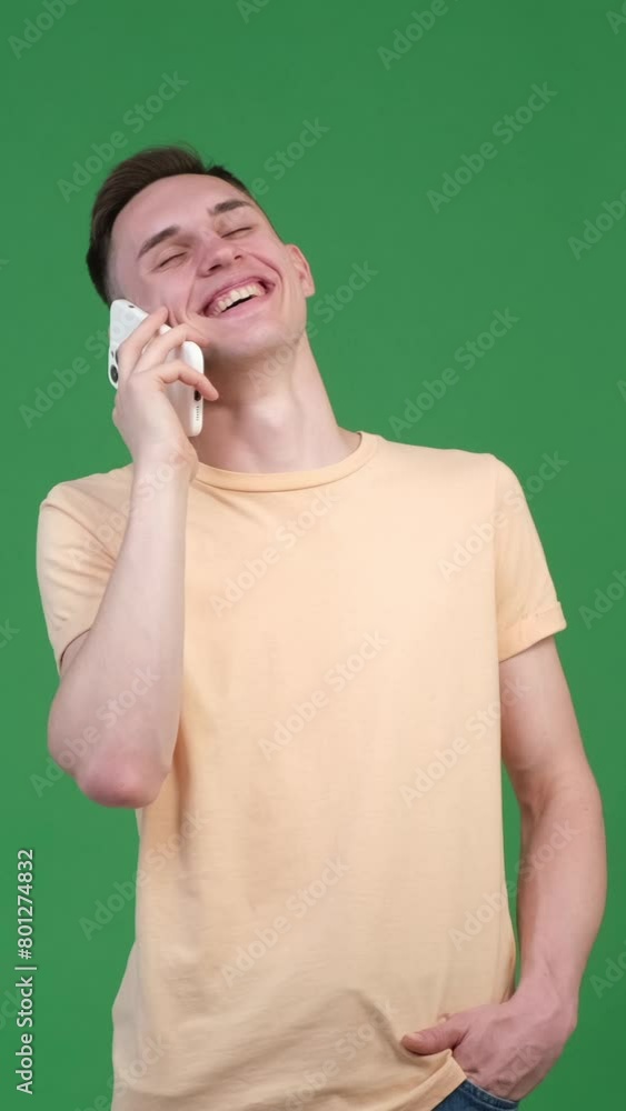 Cheerful Caucasian young adult man talking on call using smartphone and laughing on green background. Remote communication. Vertical video. Cheerful Caucasian young adult man talking on call using smartphone and laughing on green background. Remote communication. Vertical video.