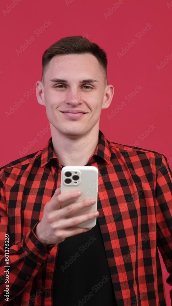 Portrait of a friendly, relaxed Caucasian man, student or businessman, holding mobile phone and looking at camera on red background. Vertical video. Portrait of a friendly, relaxed Caucasian man, student or businessman, holding mobile phone and looking at camera on red background. Vertical video.
