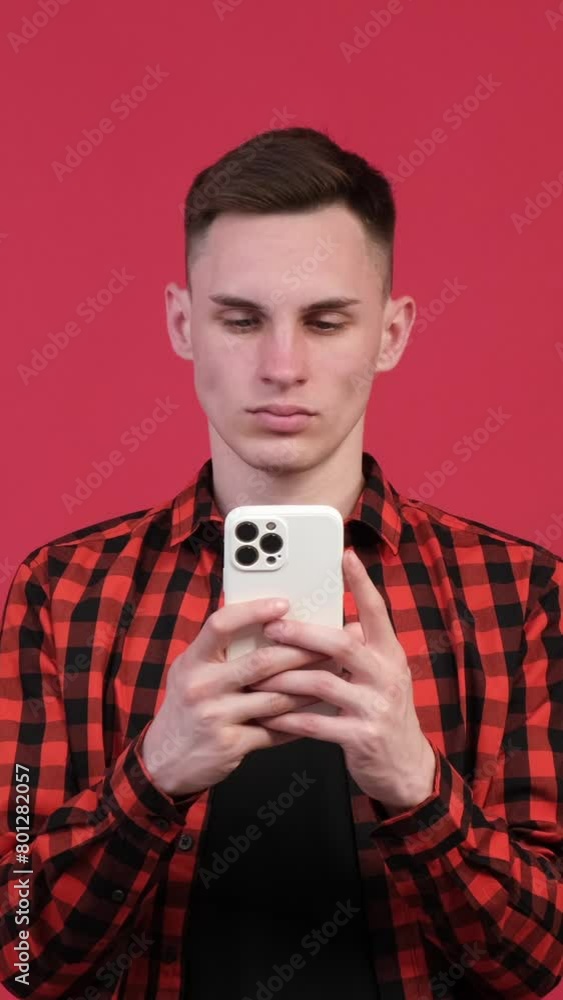 Serious Caucasian man with smartphone chatting online or surfing internet on red background. Social media and remote entertainment. Vertical video. Serious Caucasian man with smartphone chatting online or surfing internet on red background. Social media and remote entertainment. Vertical video.