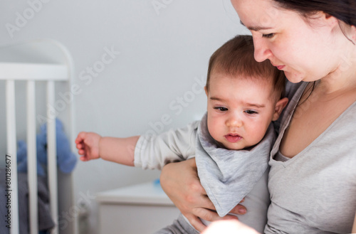 Exhausted baby sitting on the lap of his mother. Portrait of depressed woman sitting with her tired little child on bed at home, feeling baby blues, headache or migraine. Baby has a fever.