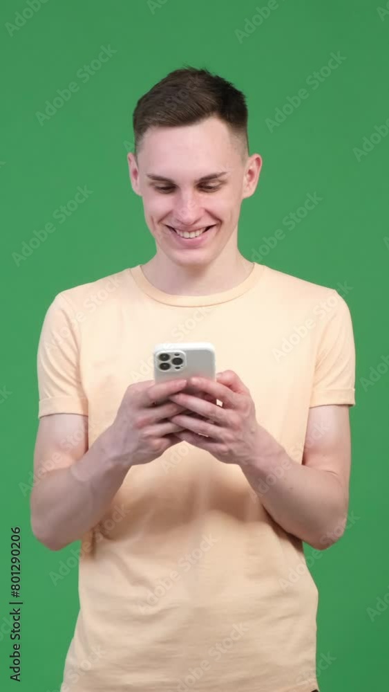 Positive and cheerful Caucasian young man resting with mobile phone, spending leisure time with online entertainment on green background. Vertical video. Positive and cheerful Caucasian young man resting with mobile phone, spending leisure time with online entertainment on green background. Vertical video.