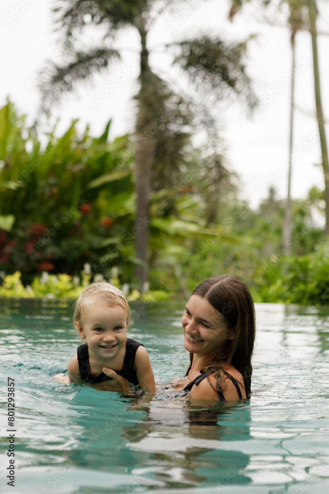 Happy family - mother, baby daughter learn to swim. Joyful kid has fun in luxury resort swimming pool. Travel lifestyle at summer beach vacation, children water sports activity, swimming lesson Happy family - mother, baby daughter learn to swim. Joyful kid has fun in luxury resort swimming pool. Travel lifestyle at summer beach vacation, children water sports activity, swimming lesson