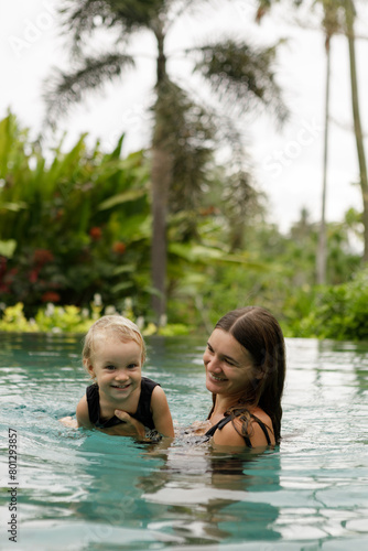 Happy family - mother, baby daughter  learn to swim. Joyful kid has fun in luxury resort swimming pool. Travel lifestyle at summer beach vacation, children water sports activity, swimming lesson  © Yevhenii