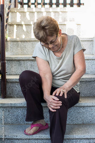 Senior woman holding the knee with pain. Senior woman sitting on stairs and suffering from pain in leg at home. Elderly woman suffering from pain in knee at home.