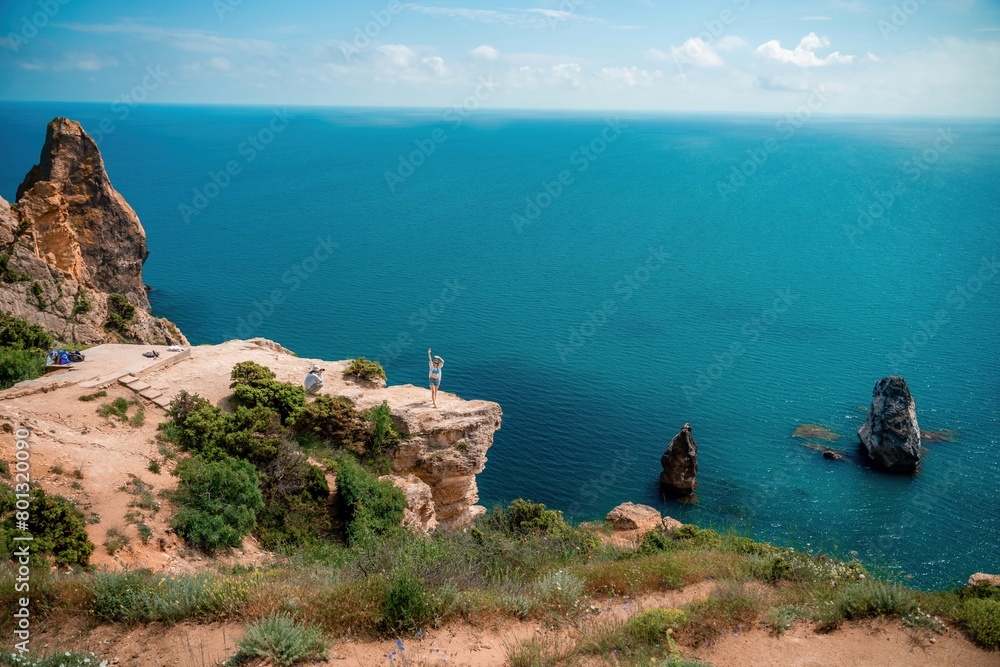 A woman stands on a cliff overlooking the ocean. The water is blue and the sky is clear.