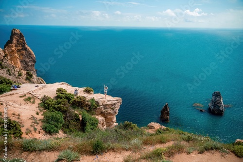 A woman stands on a cliff overlooking the ocean. The water is blue and the sky is clear. © svetograph