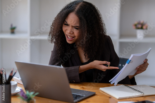 Stressed African American businesswoman reviewing documents while working on a laptop in a modern office.