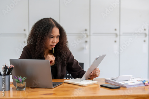 Stressed African American businesswoman reviewing documents while working on a laptop in a modern office.