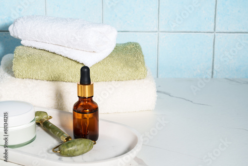 Skin care products in the bathroom. Face cream, serum bottle, jade roller and stack of towels.