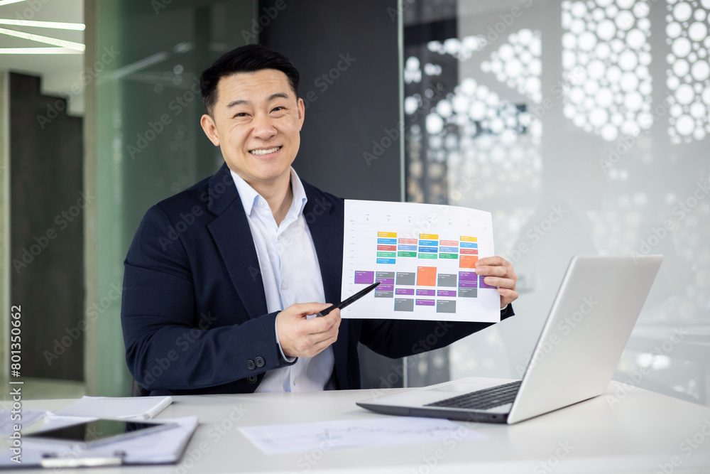 A cheerful Asian businessman in a suit presents a colorful project timeline in a modern office, demonstrating professionalism and organization.