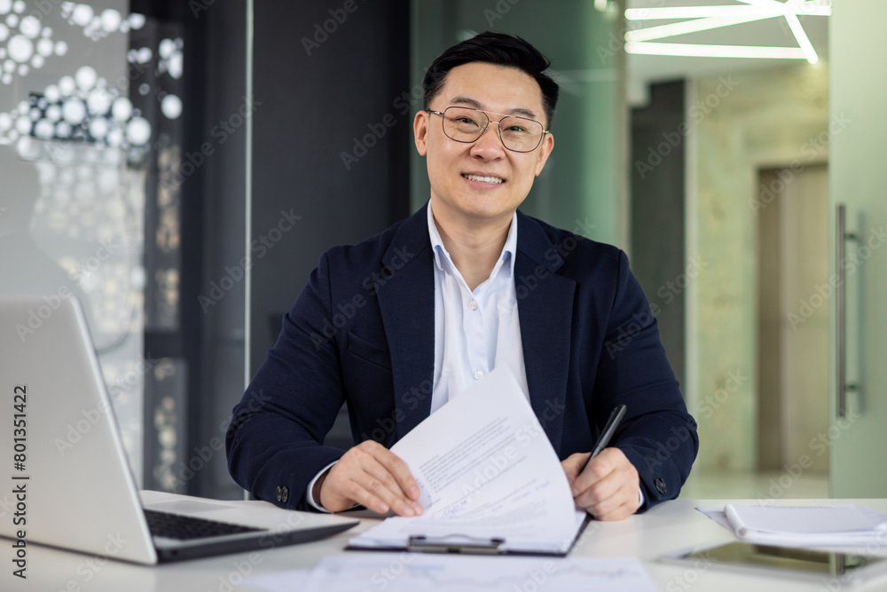 Confident Asian businessman smiling while working at a modern office desk, reviewing documents with laptop and pen in hand. Confident Asian businessman smiling while working at a modern office desk, reviewing documents with laptop and pen in hand.