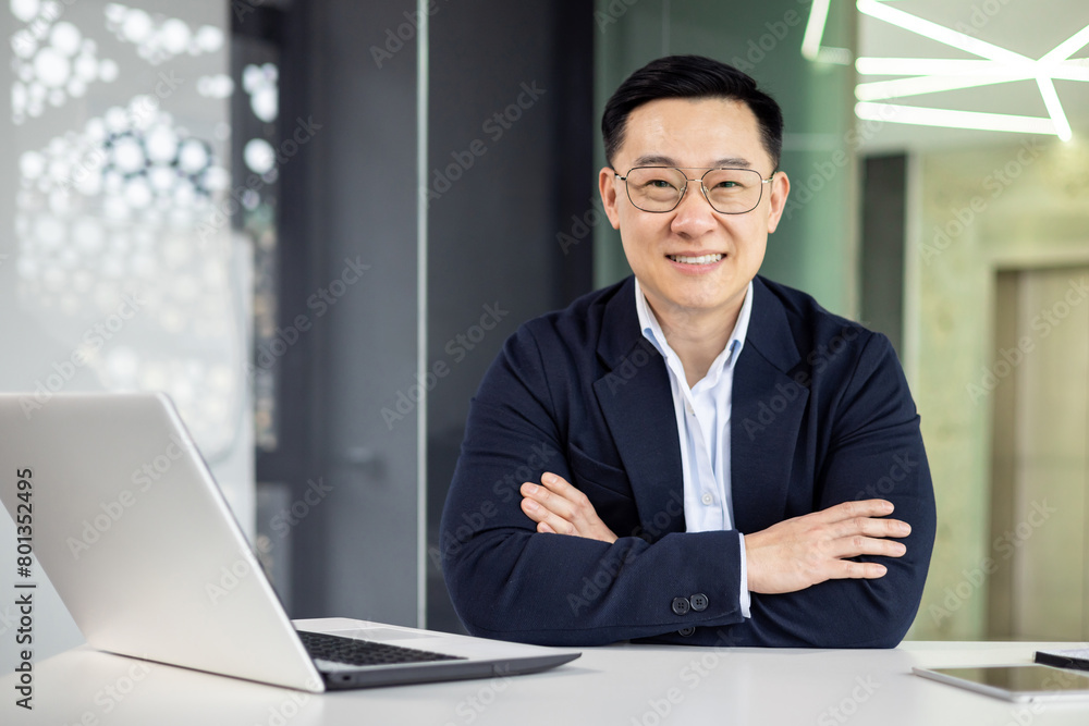 A well-dressed Asian businessman smiles confidently while sitting at his modern office desk, portraying professionalism and success in a corporate environment. A well-dressed Asian businessman smiles confidently while sitting at his modern office desk, portraying professionalism and success in a corporate environment.