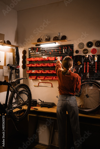 Rear view of young woman standing near wall with tools in workshop. Bicycle repair and maintenance concept.