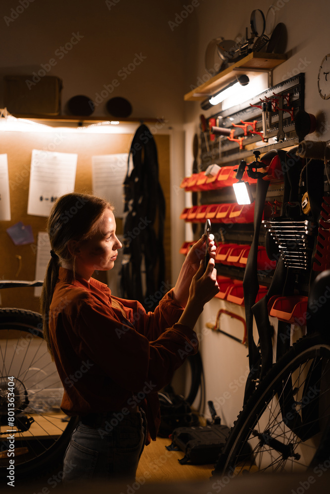 Obraz premium Female mechanic repairing bicycle in workshop, woman standing near wall with tools in garage. Bicycle maintenance concept.