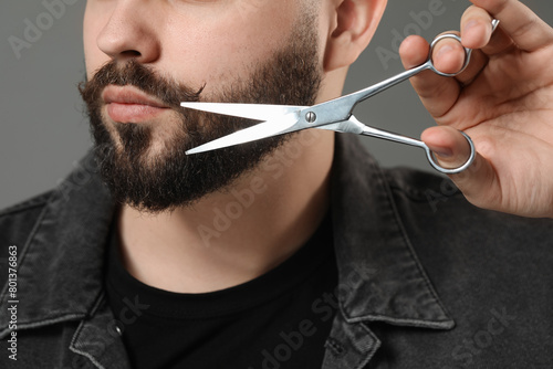 Handsome young man trimming beard with scissors on grey background, closeup