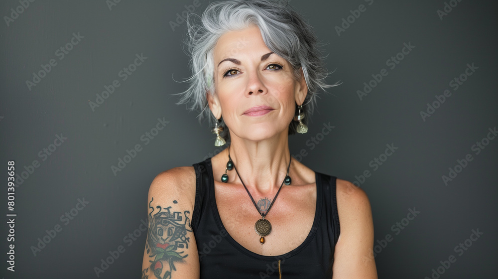 Naklejka premium Captivating photo of an attractive middle-aged woman with grey hair and tattoos, wearing earrings, necklace, and a black tank top.