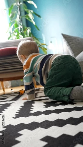 Back view of the unrecognizable boy in casual clothes, craws on floor and playing car toys while spending time in cozy living room at home Vertical footage. photo