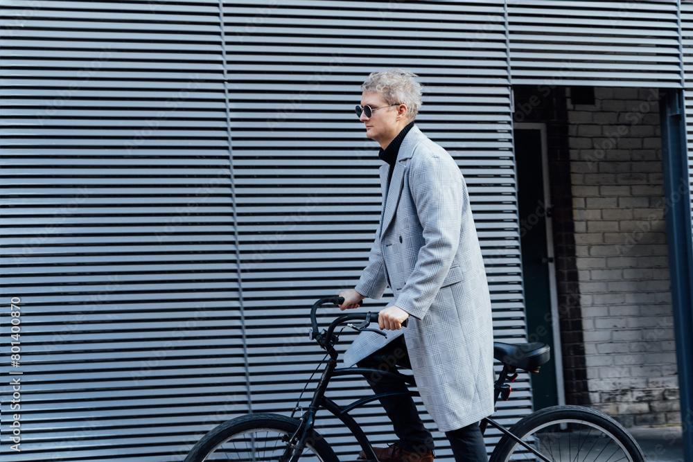 Stylish male in checked coat and sunglasses riding on retro bicycle near gray urban wall in sunny day. Neutral carbon footprint transportation. Green eco friendly mobility sustainable transport Stylish male in checked coat and sunglasses riding on retro bicycle near gray urban wall in sunny day. Neutral carbon footprint transportation. Green eco friendly mobility sustainable transport