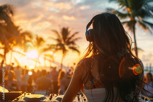 A young woman DJ playing music at sunset on the beach as a crowd dances. It was a summer party at an outdoor club or bar on a tropical island. The concept was a summer vacation nightclub scene