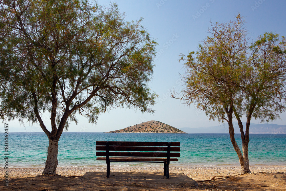 A wooden bench and two pine trees by the sea, during a sunny summer day, contemplating a small triangular shaped islet the Saronic gulf, in Salamina island, Attica region, Greece, Europe. A wooden bench and two pine trees by the sea, during a sunny summer day, contemplating a small triangular shaped islet the Saronic gulf, in Salamina island, Attica region, Greece, Europe.