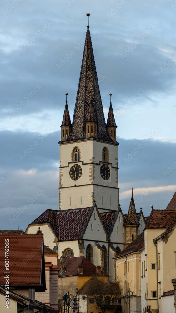 The clock tower is located in the center of the city. The tower is tall with a long, octagonal spire topped with a weather vane. The spire is covered with green glazed tiles that shine in the sunlight