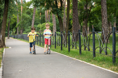 Two attractive European boys brothers, wearing red and white checkered shirts, standing on scooters in the park. They laughing, smiling, hugging and having fun. Active leisure time with kids