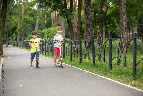 Two attractive European boys brothers, wearing red and white checkered shirts, standing on scooters in the park. They laughing, smiling, hugging and having fun. Active leisure time with kids
