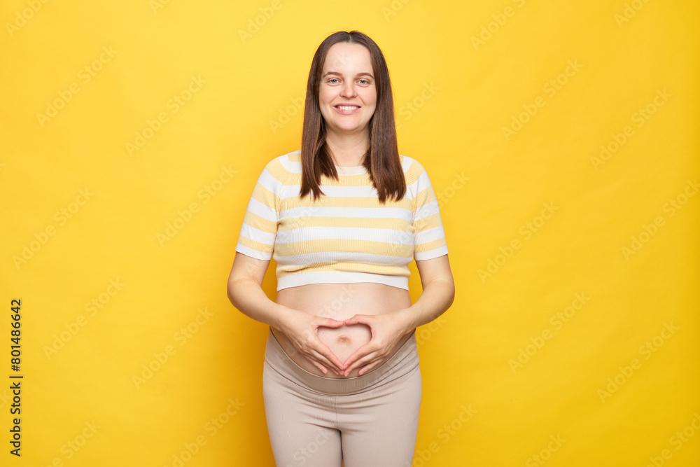 Calm positive optimistic Caucasian pregnant woman with bare belly wearing casual top isolated over yellow background making heart shape with hands expressing love to her future baby Calm positive optimistic Caucasian pregnant woman with bare belly wearing casual top isolated over yellow background making heart shape with hands expressing love to her future baby