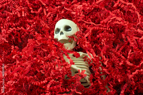 Human skeleton with skull partially buried in red crinkled shredded paper background